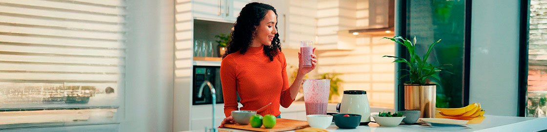 foto-de-mujer-desayunando-en-su-cocina-tomando-un-licuado-frente-a-un-plato-de-avena-con-manzanas-y-plátanos-sobre-la-barra