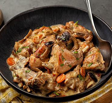 Foto de un plato con conejo guisado, junto a una cuchara sobre una tabla de madera.