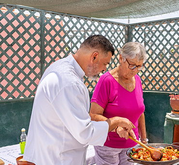 Foto de un señor y una señora cocinando paella en la terraza de una casa.