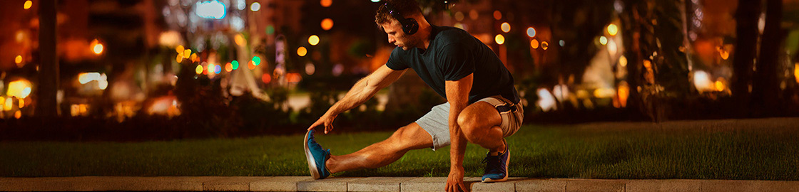 Foto de joven en ropa deportiva y con audífonos calentando en un parque por la noche antes de empezar a ejercitarse