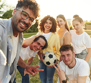 Foto tipo selfie de un grupo de amigos en el parque, con una portería de fondo, y una de ellas sostiene un balón de fútbol.