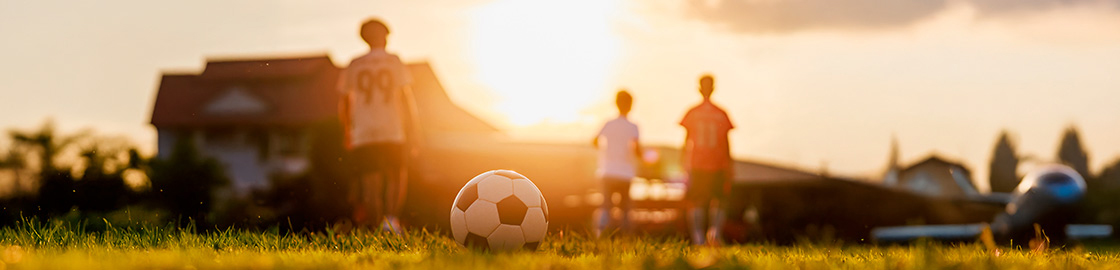 Foto de un balón de fútbol sobre el pasto en primer plano y en segundo plano 3 niños con jerseys de equipos de fútbol.