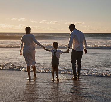 Foto de una familia, papá, hija y mamá; tomados de manos a orillas del mar, de espaldas a la cámara.