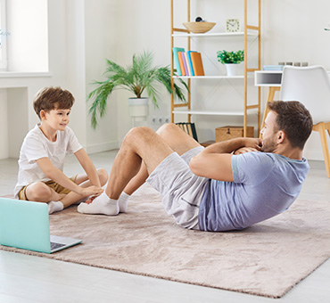 Foto de un papá atlético haciendo abdominales en la sala de su casa en compañía de su hijo.