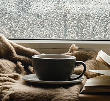 Foto desde dentro de una habitación donde se ve que afuera está lloviendo por la ventana mojada y dentro vemos una cobija, un té y un libro.