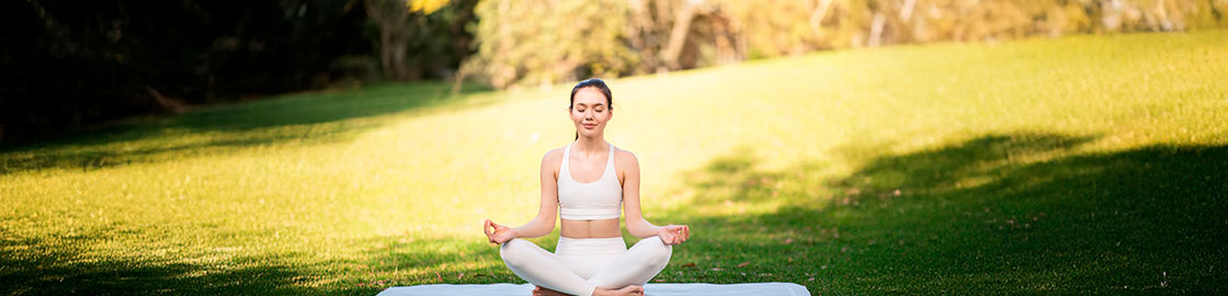 Foto de una mujer joven con ropa deportiva, sentada de piernas cruzadas sobre un tapete de yoga en el parque con los ojos cerrados y rostro concentrado.