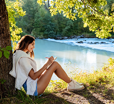 Foto de una mujer joven, sentada a la orilla de un lago con la pared recostada en un árbol, escribiendo en su diario con una sonrisa en su rostro