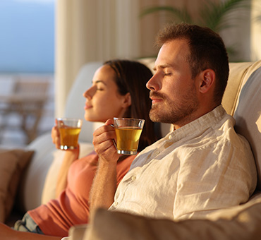 Foto de un hombre y una mujer sentados en el sillón de la salsa, cada uno sosteniendo una taza de té con los ojos cerrados y expresión relajada, disfrutando el aroma del té