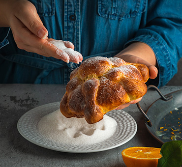 Foto de las manos de una mujer bañado de azúcar un pan de muerto sobre la barra de una cocina